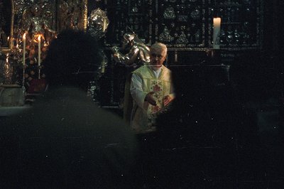 A dimly lit scene depicts a man in a white ecclesiastical robe administering communion. He holds a chalice, facing a partiall...