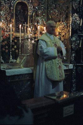 A Roman Catholic priest, wearing ornate vestments, stands before an elaborately carved and gilded altar, illuminated by numer...