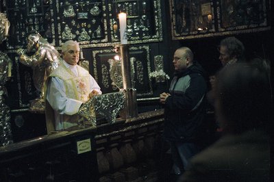 A Roman Catholic priest in vestments stands before a silver reliquary, appearing to perform a ritual. Two men observe from be...