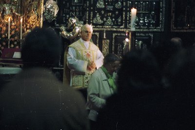 A Catholic priest in vestments elevates a host during a ceremony. Intricate, gilded altar decorations and flickering candles ...