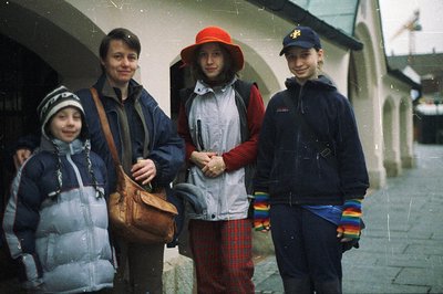 A family portrait showcasing outdoor winter wear. Four individuals stand in a covered arcade, likely in an alpine village. Vi...