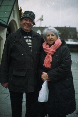 A smiling couple stands outdoors, appearing to be on vacation. The man wears a newsboy cap and dark coat; the woman, a pink s...