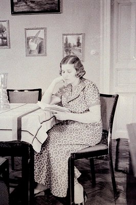 A woman seated at a table, engrossed in reading. She wears a patterned, long-sleeved dress and appears contemplative. A patte...