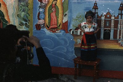 A young girl in traditional dress poses on a small stool before a mural depicting the Virgin Mary and a church facade. Likely...