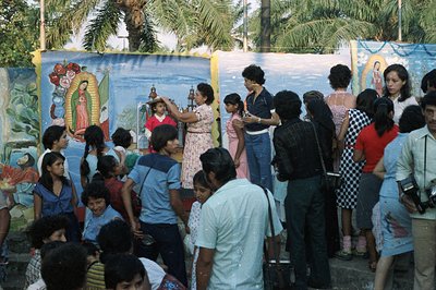 A vibrant scene: a crowd gathers before murals of the Virgin Mary. A woman in a floral dress raises a religious icon, while o...
