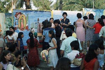 A vibrant outdoor gathering features a large mural of the Virgin of Guadalupe. People in 1970s attire—floral dresses, bell-bo...