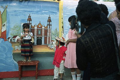 A young girl in traditional Mexican folk dress poses in front of a painted mural depicting a church, likely for a staged phot...