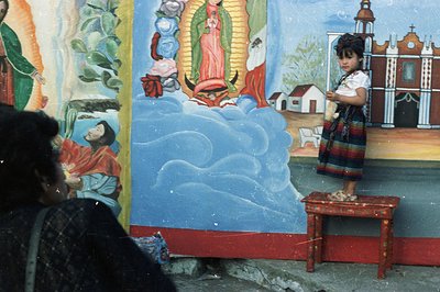 A young girl in traditional clothing stands on a small table before a vibrant mural depicting the Virgin Mary and a church fa...