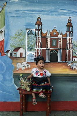 A young girl in traditional Mexican folk dress sits perched on a vibrant, hand-painted mural depicting a church and townscape...