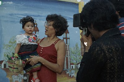 A young child in traditional clothing is held by a woman in a red dress, posing in front of a painted backdrop resembling a E...