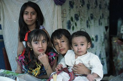 A poignant portrait of three children, seemingly indigenous, set against a patterned fabric backdrop. A young girl in a vibra...