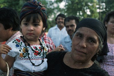 A young girl in traditional embroidered clothing rests on a woman’s shoulders. The woman wears a dark headscarf. Background f...
