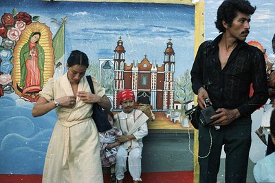 A woman in a traditional dress adjusts a boy’s hat while he sits on a pedestal before a vibrant mural depicting the Virgin of...