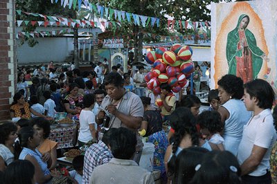Crowded outdoor gathering features a large image of the Virgin of Guadalupe. Festive decorations, balloons, and food stalls i...