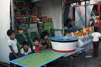A vibrant street scene captures children near a vendor's stall overflowing with colorful plastic containers. The stall displa...