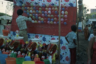 A vibrant, makeshift carnival game display featuring numerous balloons affixed to a floral-patterned backdrop. Two figures as...