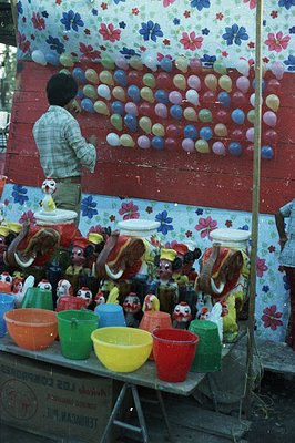 A vendor stands near a colorful display of ceramic rooster figurines and plastic buckets, set against a floral-patterned back...