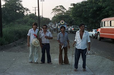 Four men stand on a paved road, appearing casually posed. They wear 1970s-era fashion—bell-bottoms, open shirts, and wide col...