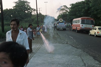 A roadside scene depicts men lighting fireworks on an asphalt road. Children and onlookers watch as the smoke rises. A bus, t...