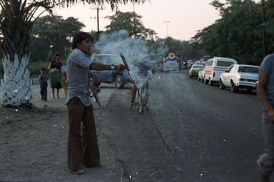 A man in flared trousers and a button-down shirt ignites a string of firecrackers on a dusty road. A van, several cars, and o...