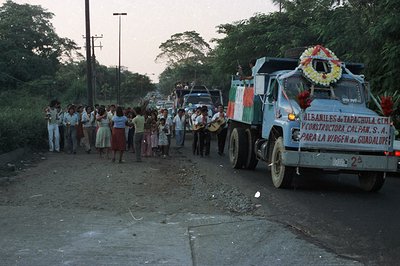 A celebratory procession unfolds on a rural road. A light blue dump truck, adorned with flowers, leads a group of people, som...