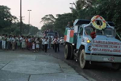 A parade featuring a decorated dump truck and a marching band moves down a road lined with onlookers. The truck bears signage...