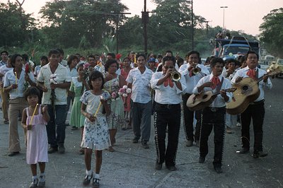 Procession featuring a musical ensemble and community members in traditional attire. Candle bearers lead the group, followed ...