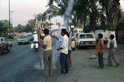 Four men stand on a paved road, one lighting a large, smoking bundle of dried herbs or branches. Palm trees line the street, ...