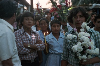 A community gathers during a religious procession. Individuals hold candles and bouquets of flowers. Clothing styles and hair...