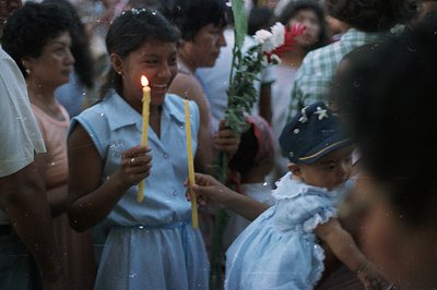 A young girl in a light blue dress and cap holds a bouquet of flowers while a teenage girl, also in blue, carries two lit can...