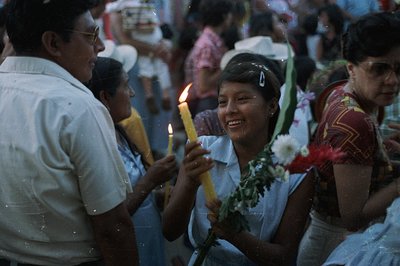 A group gathers in what appears to be a festive, outdoor scene. A young woman holds a lit candle, smiling warmly, surrounded ...