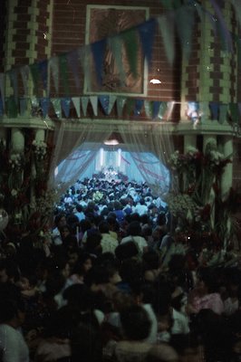 Large crowd gathers beneath decorative flags and floral arrangements, likely for a religious festival or community celebratio...