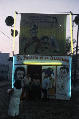 A vibrant, hand-painted sign proclaims "El Palacio de la Ilusion" above a small storefront. The sign depicts theatrical scene...