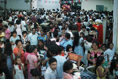 Large crowd gathered outdoors, possibly a festival or community event. People dressed in 1970s fashion—floral prints, pastel ...