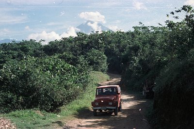 A red Fiat 500 Jeep navigates a narrow dirt road flanked by dense tropical foliage. A dormant volcano is visible in the dista...
