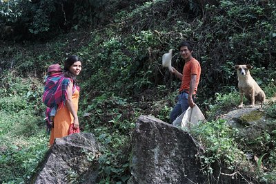 A woman carrying a child on her back stands on a rocky ledge overlooking lush vegetation. A man in an orange shirt and jeans ...