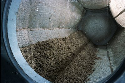Close-up image of a textile mesh container filled with what appears to be dry, granular material, possibly soil or sand. The ...
