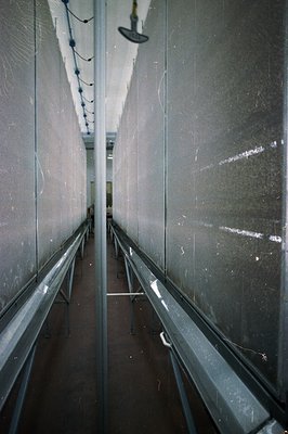 Narrow corridor features tiled walls and a metal-grated floor, illuminated by overhead fixtures. The perspective emphasizes t...