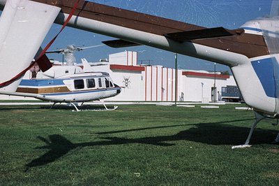 Helicopter parked on grass, partially obscured by rotor blades. Behind, a building with Art Deco-inspired red and white strip...