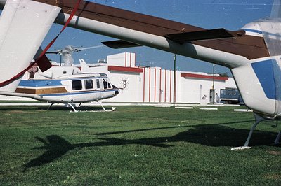 A blue and white Bell 206 helicopter sits on a grass field, partially obscured by the shadow and rotor of another helicopter....
