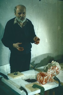 A middle-aged man with a full beard stands near a table holding a bloody, partially processed animal carcass. Likely a butche...