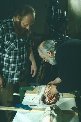 Two men examine what appears to be a sculpted or carved head form, heavily stained with red substance, likely paint or dye. T...