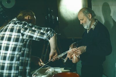 Two figures in a butcher's shop, one slicing a large cut of meat. The older man, with a white beard, is holding the meat stea...