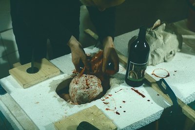 A heavily blurred image depicts a butcher's workspace with a large, blood-soaked meat mass center. A gloved hand wields a kni...