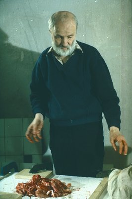 A butcher, likely middle-aged, stands behind a butcher block displaying cuts of raw meat. He wears a dark blue sweater and ap...