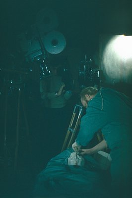 A figure in a teal work shirt is crouched, adjusting a vintage camera mounted on a tripod. Large, older studio lighting equip...