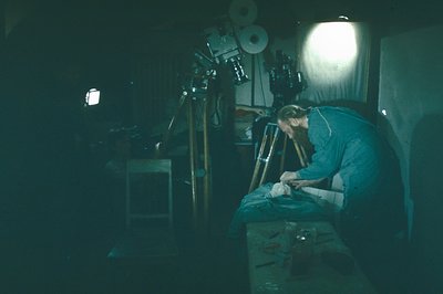 A man in a blue work shirt meticulously repairs a film reel in a dimly lit, cluttered workspace. Visible are film spools, too...