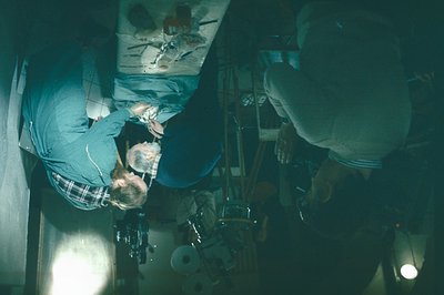 High-angle view of two figures in a workshop, likely woodworking or model-making. One individual is seated, using tools on a ...