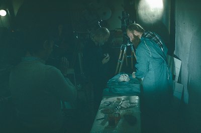 A photograph depicting three figures in a dimly lit space, likely a darkroom. Two men examine a developed photograph laid on ...