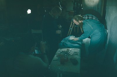 A man with a beard examines what appears to be a bird specimen on a table, while another figure observes. The scene is dimly ...